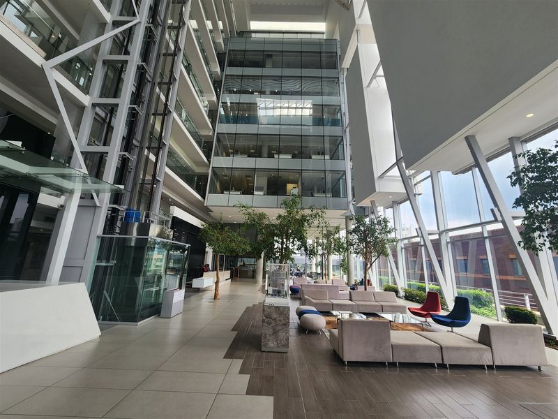 A modern, high-ceilinged office atrium featuring expansive glass curtain walls, a glass elevator, and polished tile flooring in a professional corporate environment.