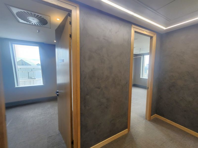 A modern office space featuring textured grey walls, carpeted flooring, recessed ceiling lighting, and integrated air conditioning vents with views through large windows.