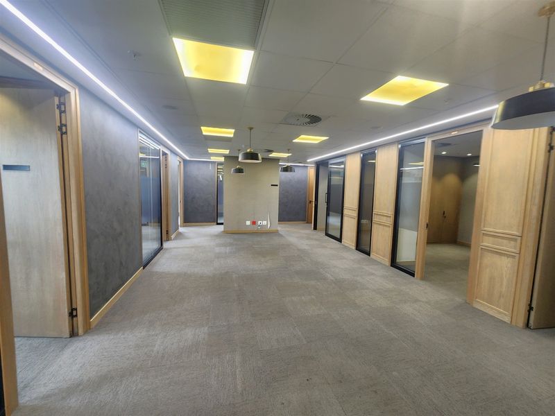 A modern, professional office corridor featuring high-quality glass partitions, timber finishes, recessed lighting, and neutral carpeting in an open, well-maintained layout.