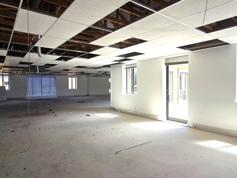 Spacious open-plan office area currently undergoing renovation, featuring exposed ceiling structure, missing tiles, and large windows providing ample natural light.