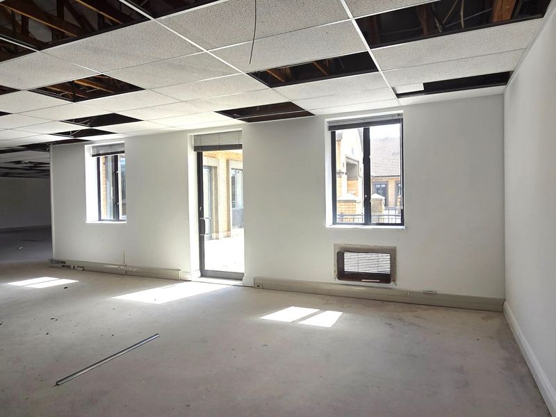 An empty office space featuring a suspended ceiling with missing tiles, large windows, and a glass door leading to an exterior area.