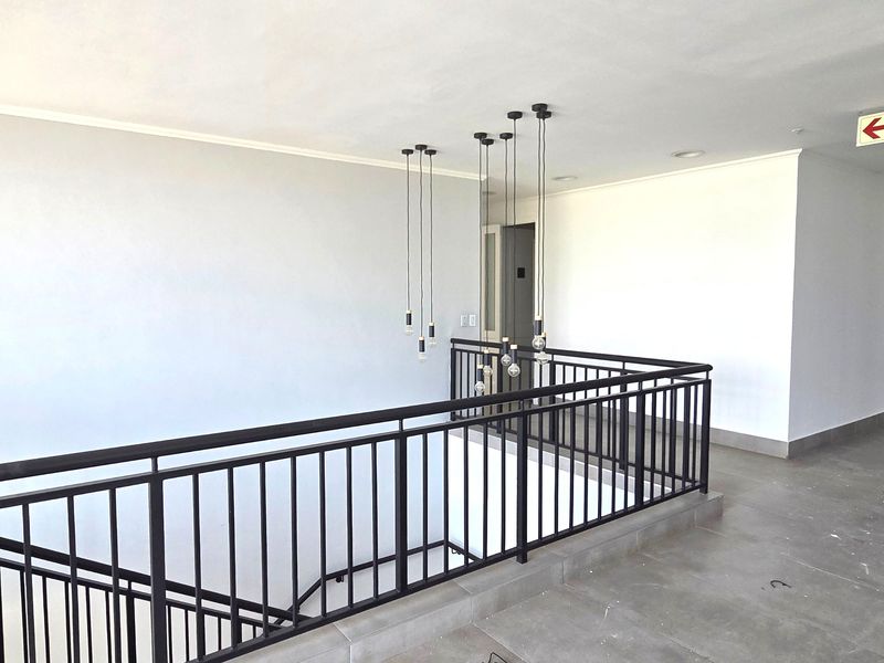 A bright and modern office hallway featuring a black metal balustrade overlooking the staircase, complemented by contemporary pendant lighting and neutral tiled flooring.