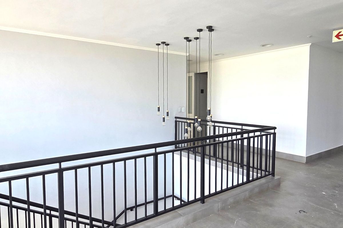 A bright and modern office hallway featuring a black metal balustrade overlooking the staircase, complemented by contemporary pendant lighting and neutral tiled flooring.