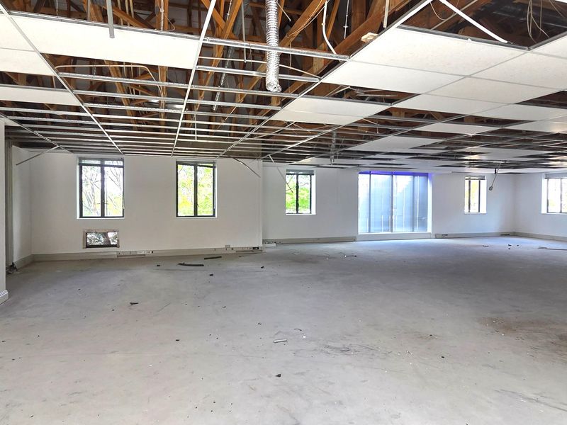 A large, open-plan office space currently undergoing renovation, featuring exposed roof trusses, a partial suspended ceiling grid, and multiple windows providing natural light.