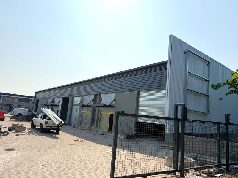 Exterior view of an industrial warehouse facility featuring corrugated metal cladding, multiple roller shutter doors, and a paved yard area under construction.