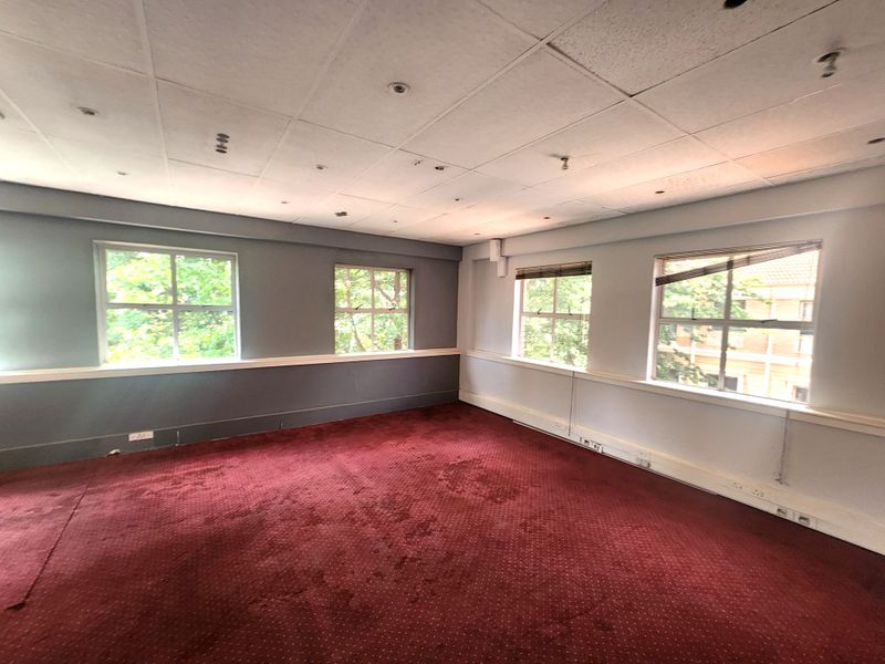 Spacious empty office area featuring a suspended ceiling, large windows providing natural light, and carpeted flooring, suitable for a professional workspace.