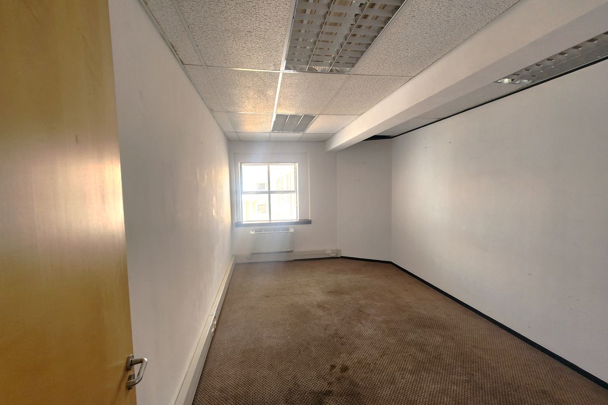 An empty office space featuring a suspended ceiling with recessed lighting, carpeted flooring, and a window-mounted air conditioning unit.