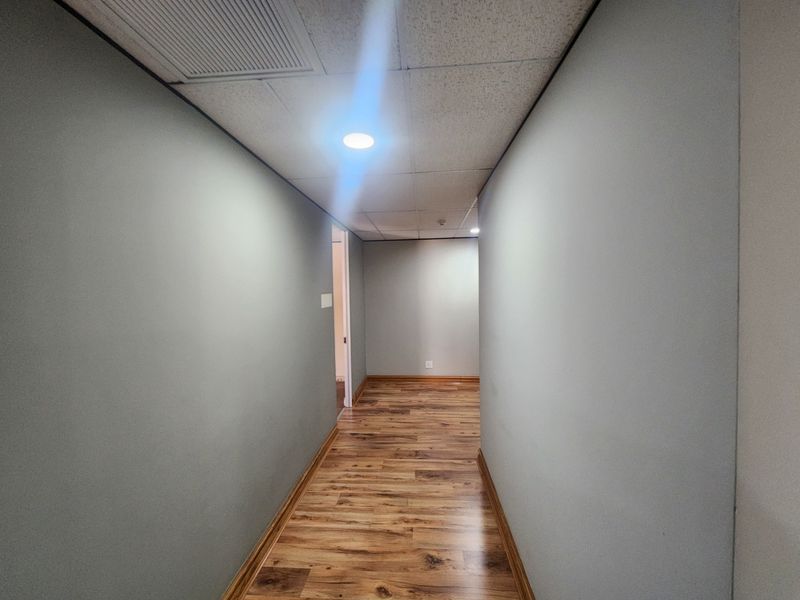 A clean, empty hallway featuring modern laminate wood flooring, a suspended ceiling with recessed lighting, and neutral grey walls.