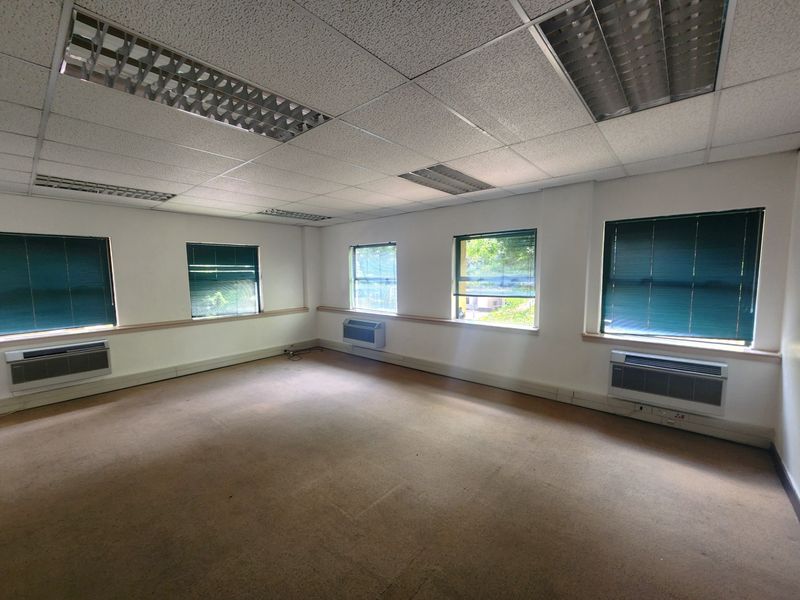 A spacious, empty office featuring suspended ceilings, recessed lighting, wall-mounted air conditioning units, and large windows with venetian blinds.