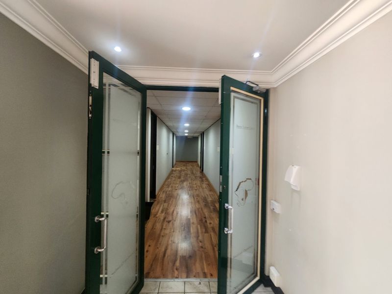 A well-lit office hallway featuring laminate wood flooring, a suspended ceiling with recessed lighting, and elegant frosted glass double doors at the entrance.