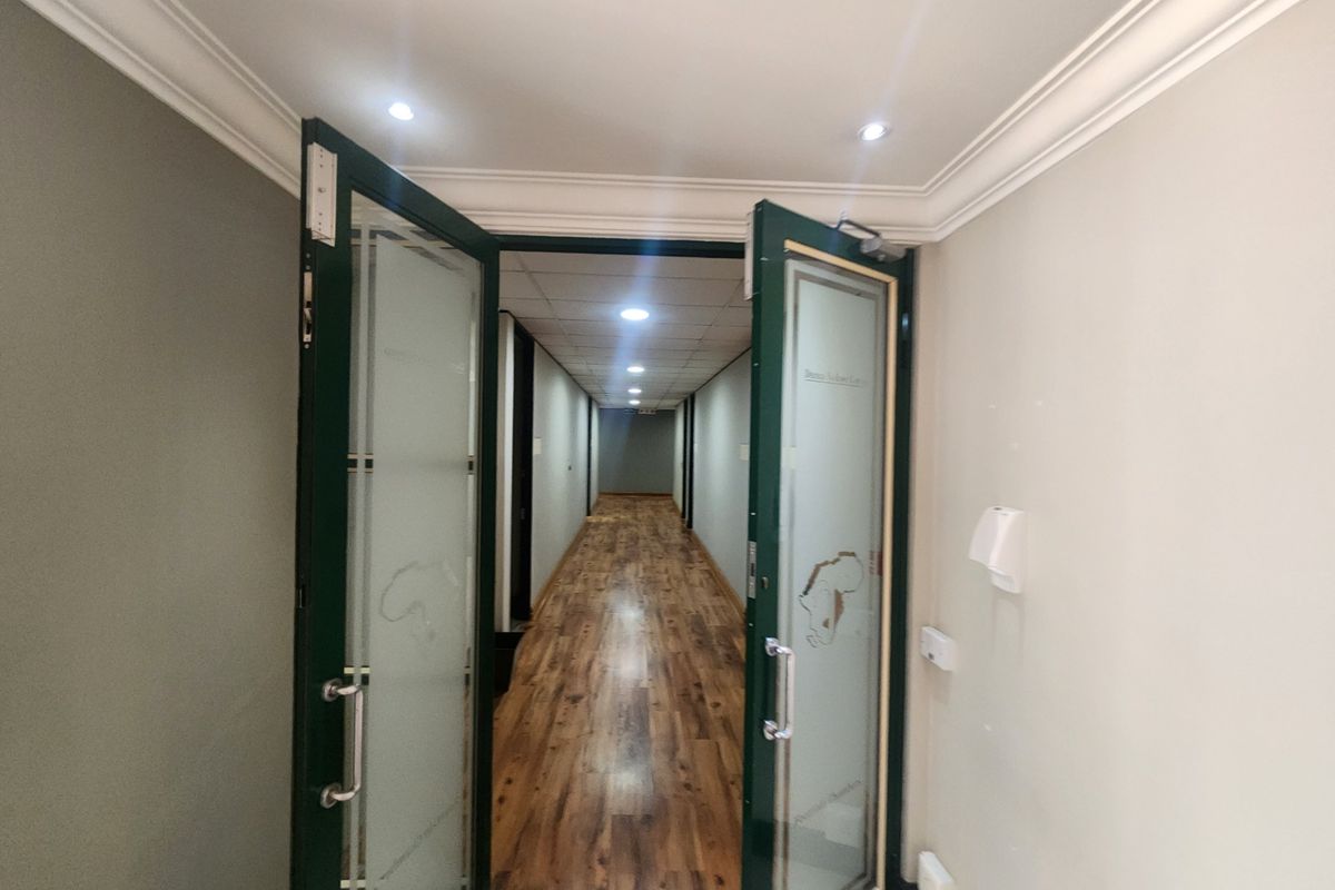 A well-lit office hallway featuring laminate wood flooring, a suspended ceiling with recessed lighting, and elegant frosted glass double doors at the entrance.