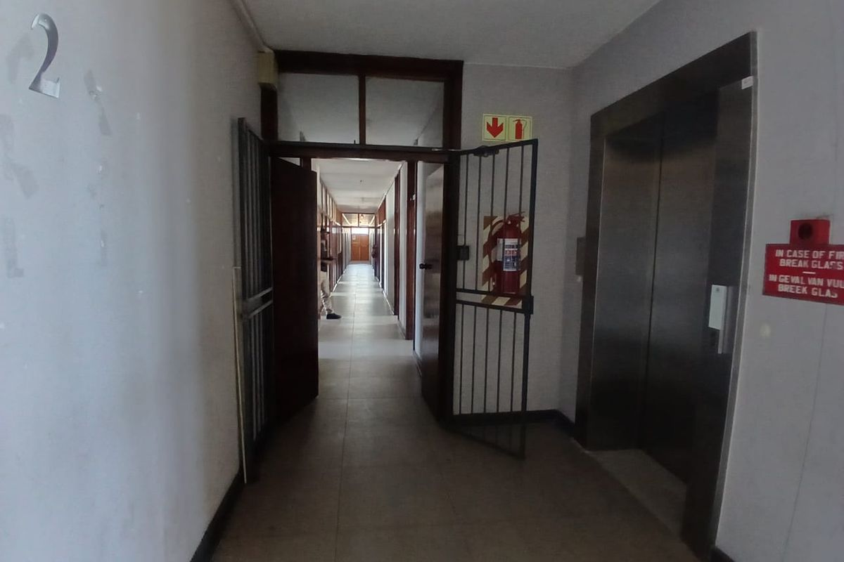 A commercial office building hallway featuring an elevator, security gate, and fire safety equipment with tiled flooring and neutral wall finishes.