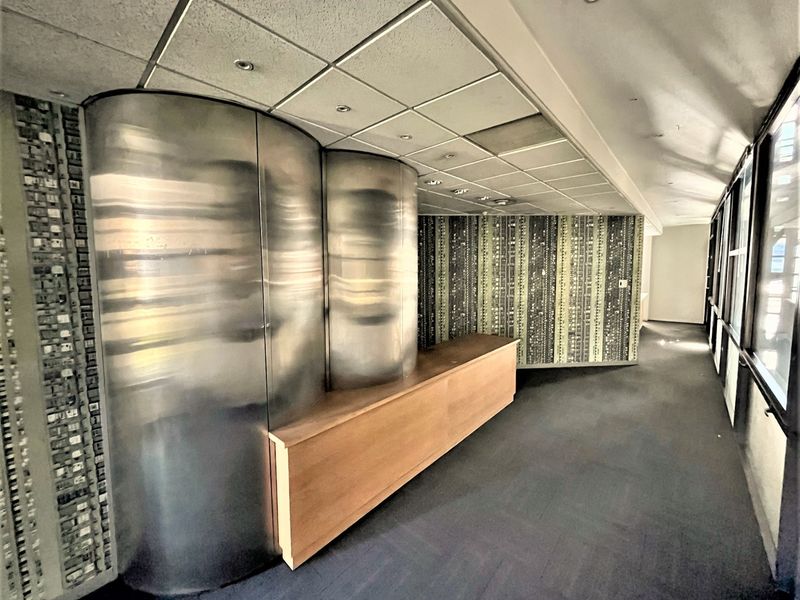 An empty office space featuring a unique curved metal reception desk, patterned wallpaper, suspended ceiling with recessed lighting, and large windows providing natural light.