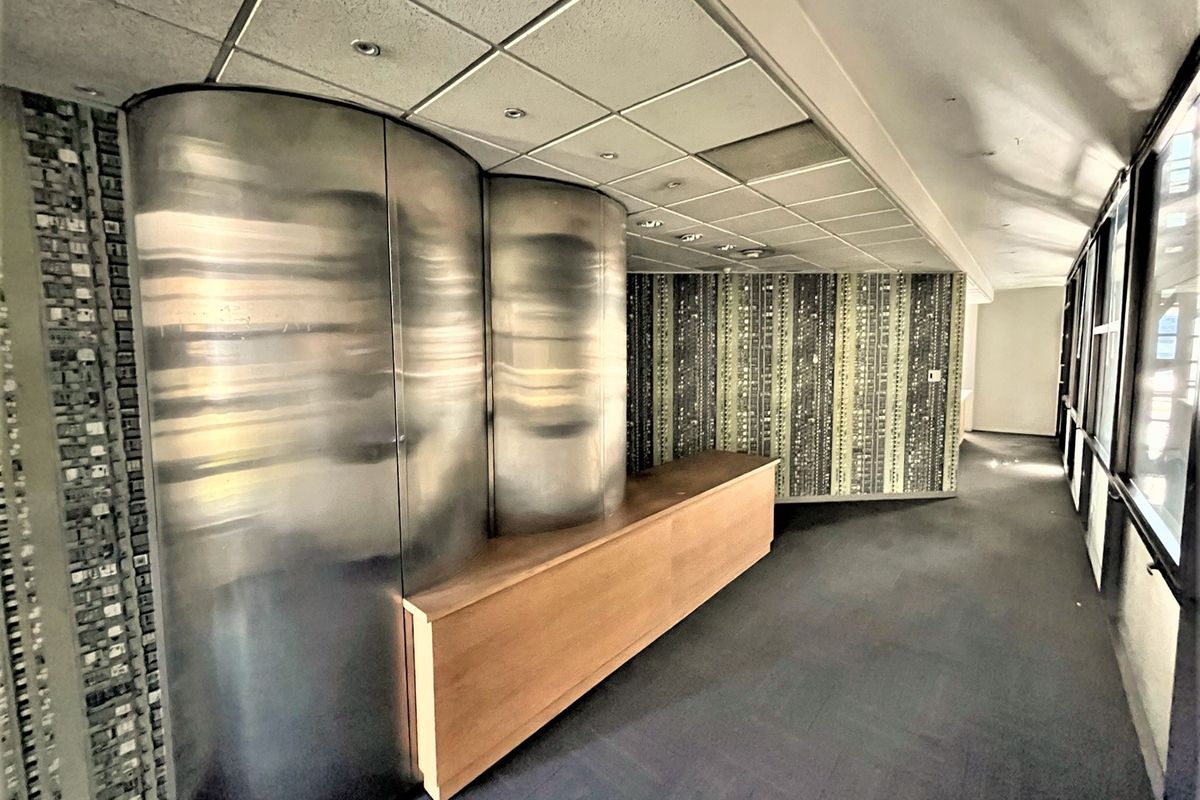 An empty office space featuring a unique curved metal reception desk, patterned wallpaper, suspended ceiling with recessed lighting, and large windows providing natural light.