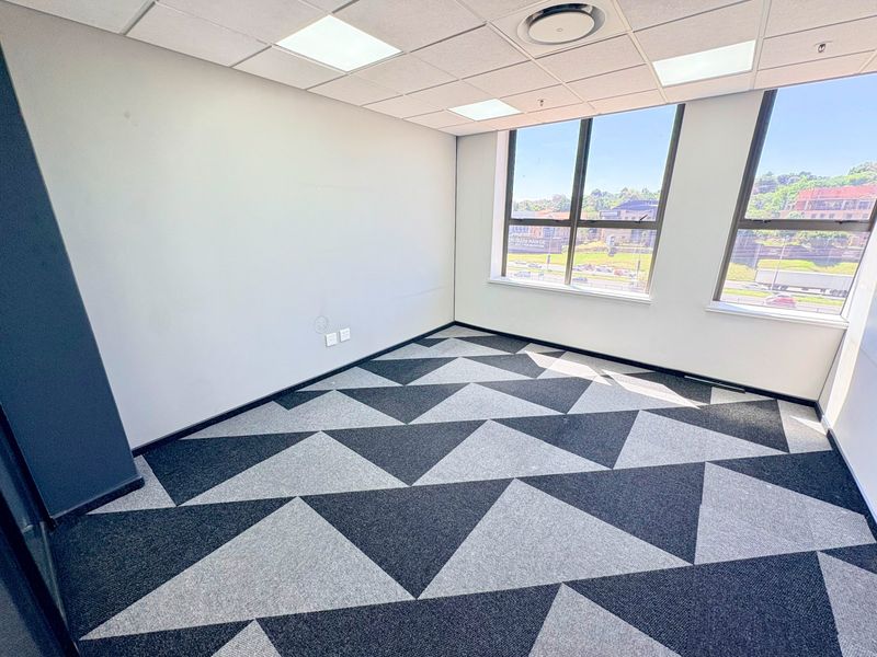 A bright, empty office space featuring modern patterned carpet tiles, a suspended ceiling with recessed lighting, and large windows providing natural light.