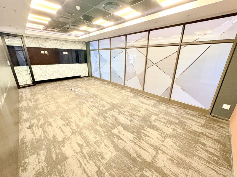 An empty office space featuring glass partition walls, carpet tiles, and a suspended ceiling with recessed lighting.