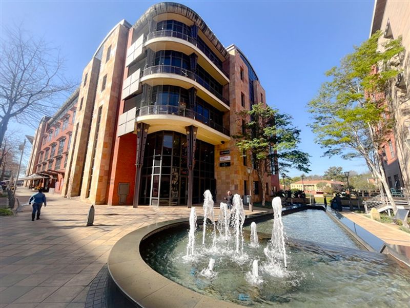 Exterior view of a modern multi-story office building featuring stone cladding, glass curtain walls, balconies, and a prominent water fountain in the foreground.