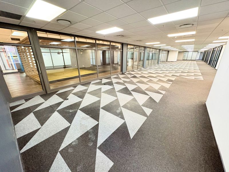 Spacious open-plan office area featuring modern glass partitions, suspended ceilings with recessed lighting, and distinctive geometric patterned carpet tiles throughout.