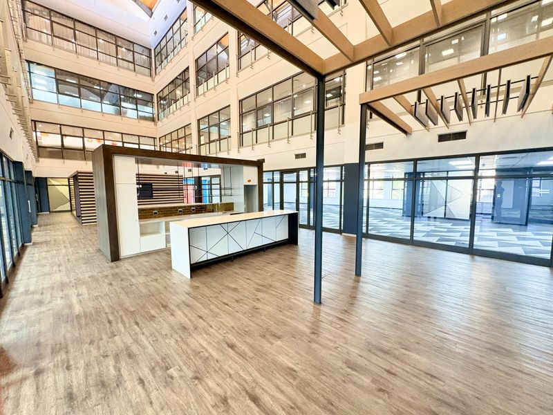 Spacious open-plan office area featuring modern wood-look flooring, a built-in kitchenette, and extensive glass partitioning within a bright, multi-level atrium setting.