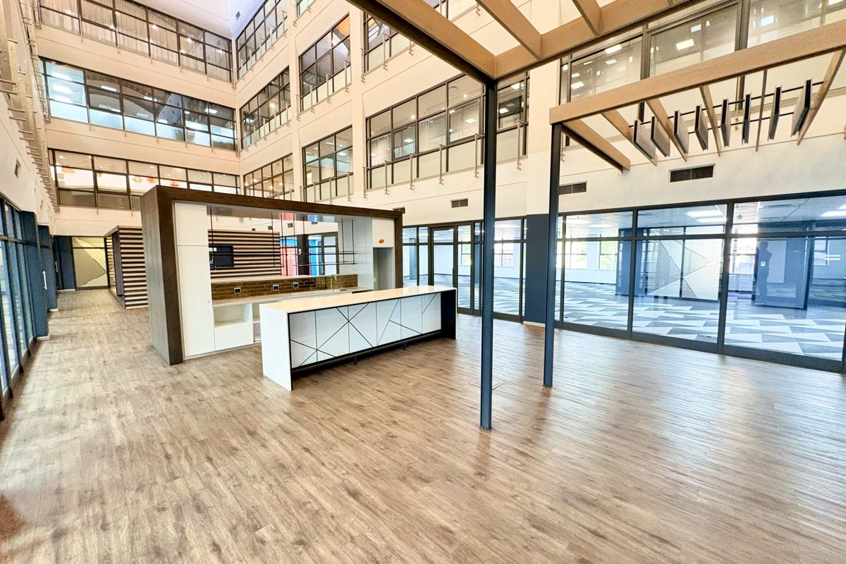 Spacious open-plan office area featuring modern wood-look flooring, a built-in kitchenette, and extensive glass partitioning within a bright, multi-level atrium setting.