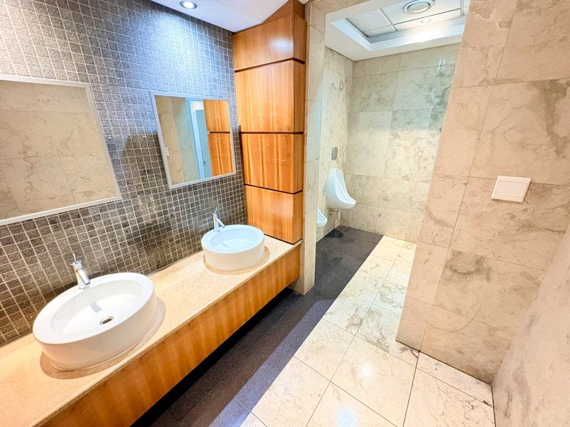 Modern office bathroom featuring a dual vessel sink vanity with stone countertop, wooden cabinetry, and tiled wall and floor finishes.