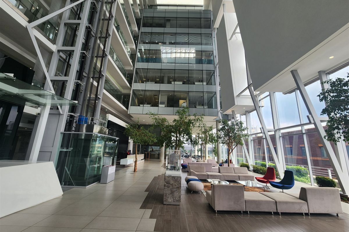 A modern, high-ceilinged office atrium featuring expansive glass curtain walls, a glass elevator, and polished tile flooring in a professional corporate environment.