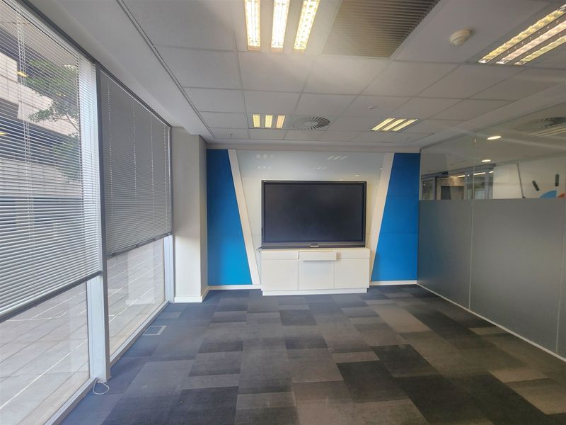 A modern, empty office space featuring carpet tiles, a glass partition wall, recessed lighting, and a built-in media cabinet with a large display screen.