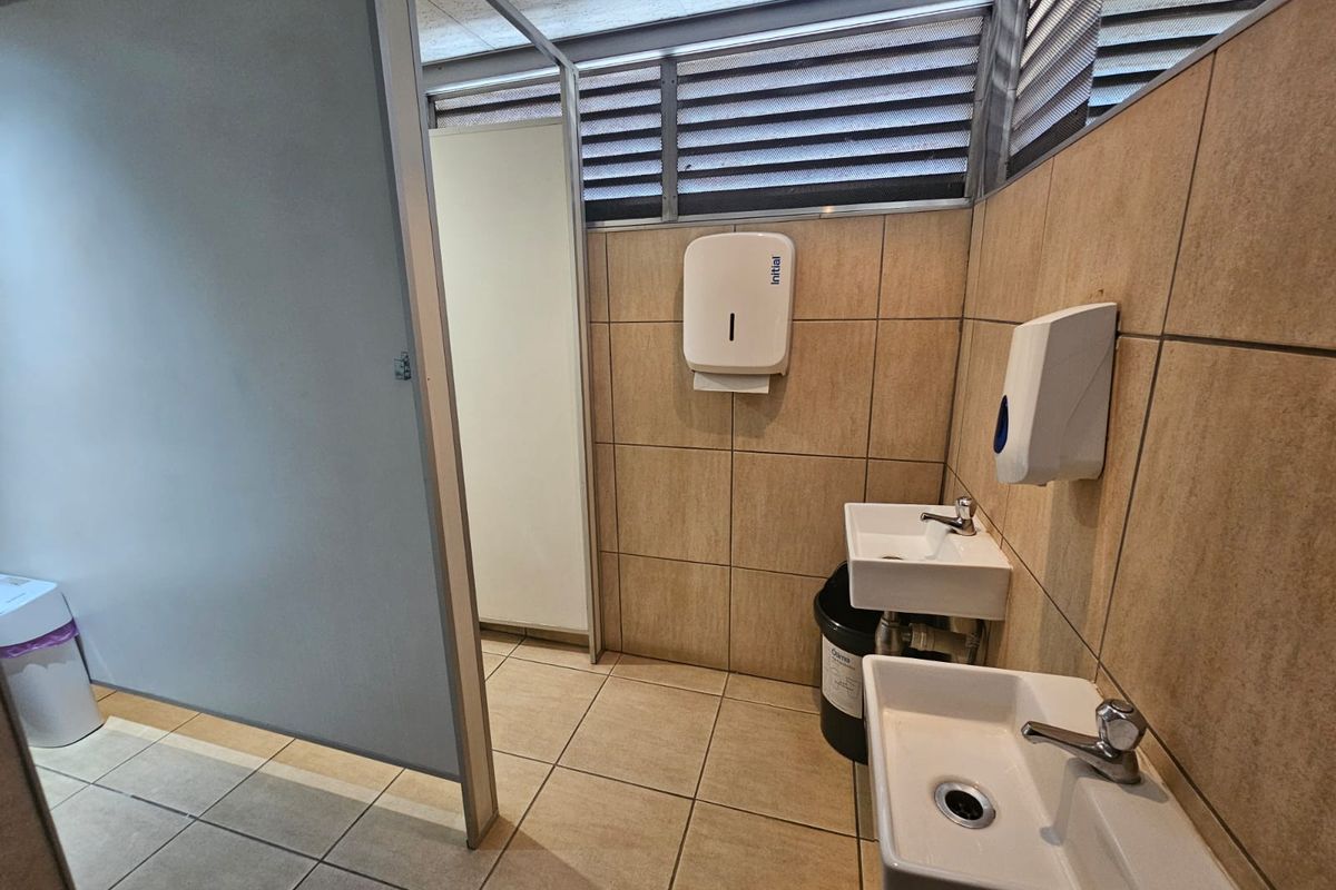 A functional office bathroom featuring dual wall-mounted basins, tiled walls and flooring, and a private partition cubicle.
