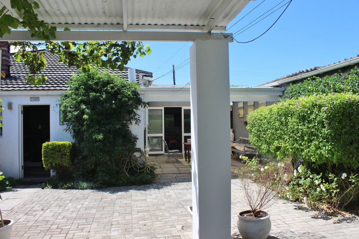 A rear exterior view featuring a paved patio area, covered walkway with a corrugated roof, and access to the building via sliding glass doors.