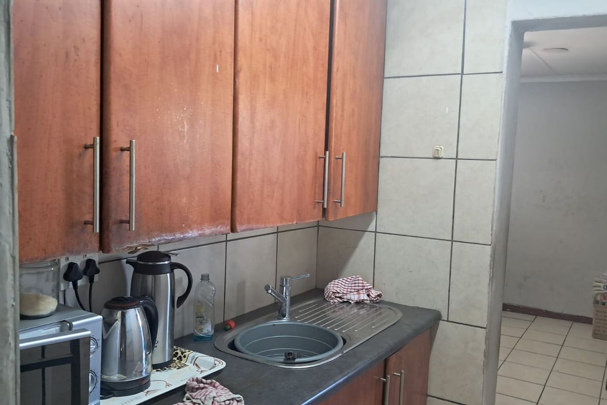 A compact kitchen area featuring wooden cabinetry, a stainless steel sink, and tiled splashbacks, leading into an adjacent utility space.