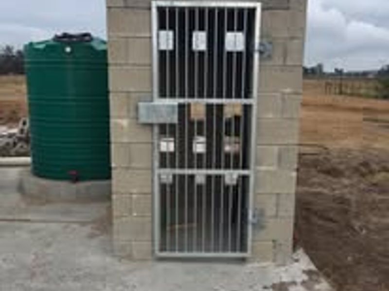 A functional utility structure featuring a concrete block enclosure with a metal security gate, accompanied by an external water storage tank on a concrete base.