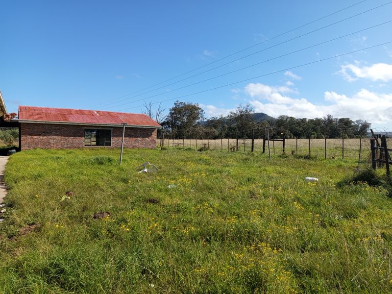 A view of the rear exterior of a face brick farm building with a corrugated metal roof, set within a large, overgrown grassy field.