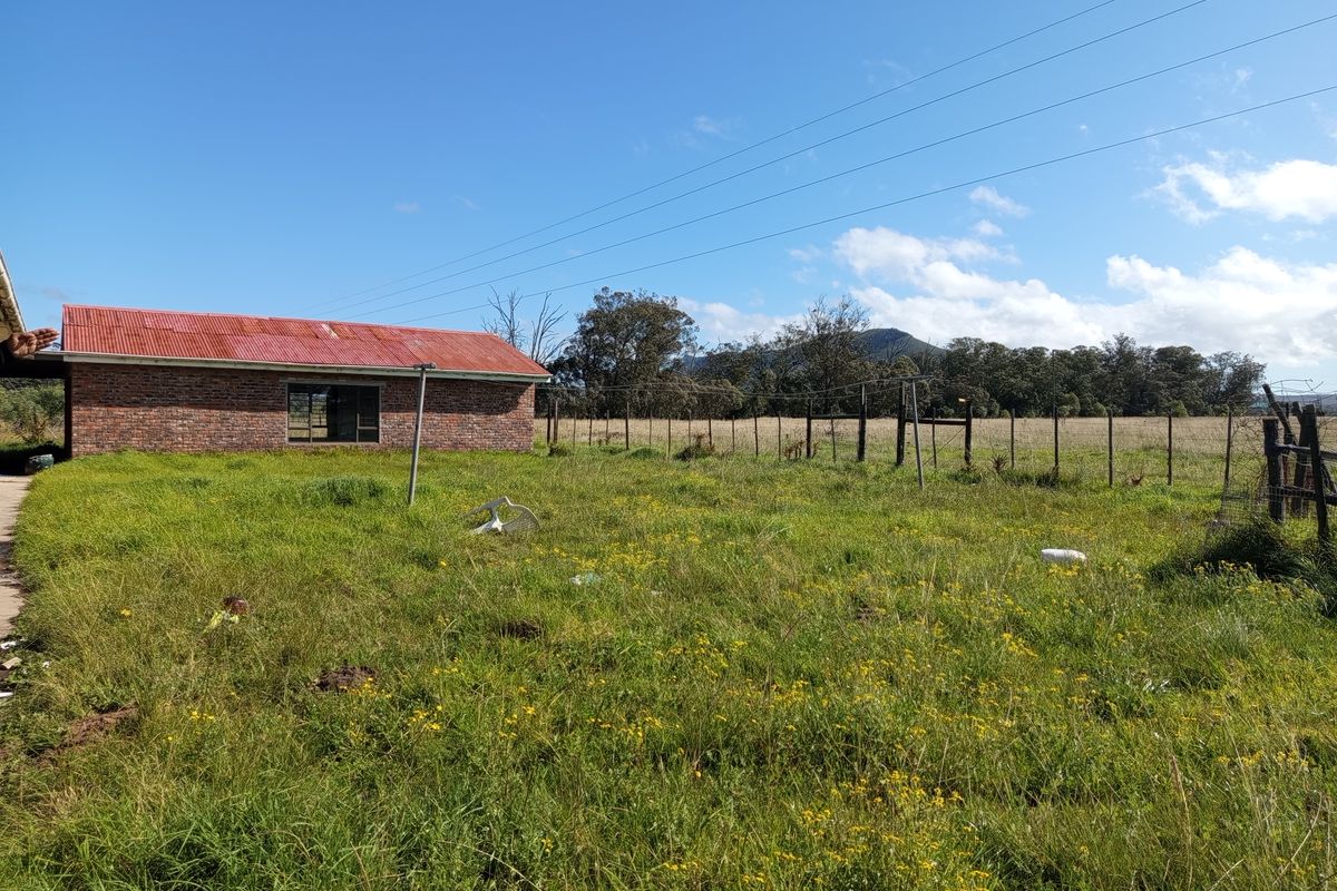A view of the rear exterior of a face brick farm building with a corrugated metal roof, set within a large, overgrown grassy field.
