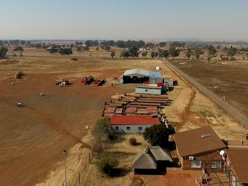 Aerial view of an industrial property featuring a large warehouse, expansive yard space, and residential buildings on a rural plot.