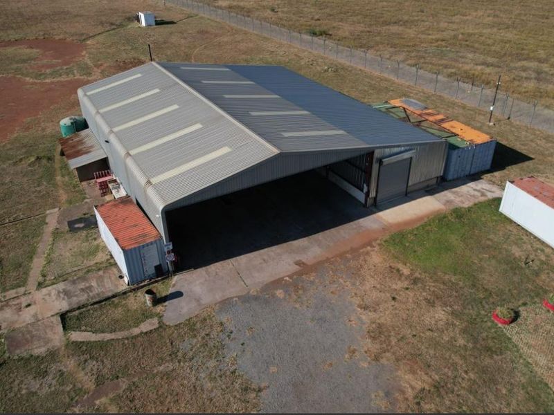 Aerial view of an industrial warehouse featuring a large corrugated metal structure, roller shutter access, and a concrete apron on a fenced plot.