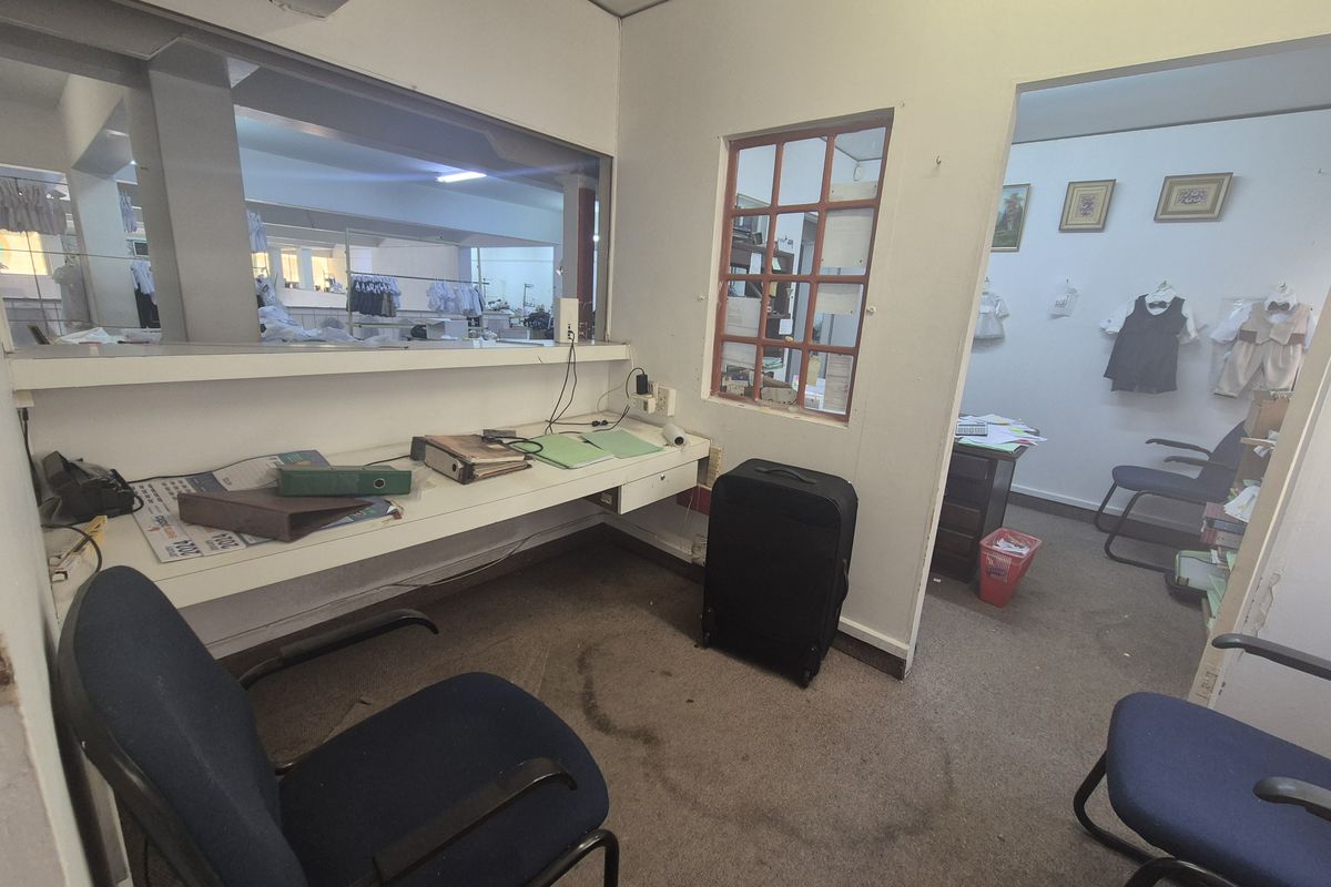 A functional office space featuring a built-in desk, internal window partition, and carpeted flooring, suitable for administrative or retail support use.
