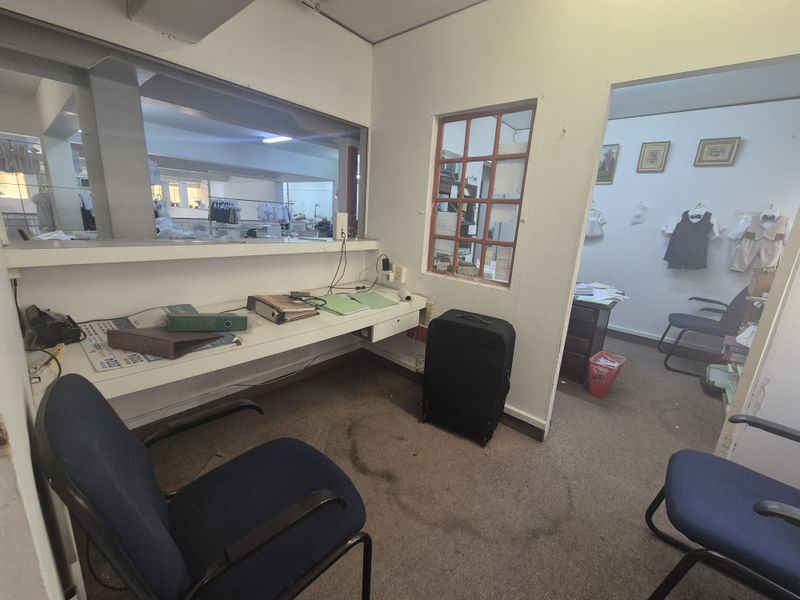 A functional office space featuring a built-in desk, internal window partition, and carpeted flooring, suitable for administrative or retail support use.