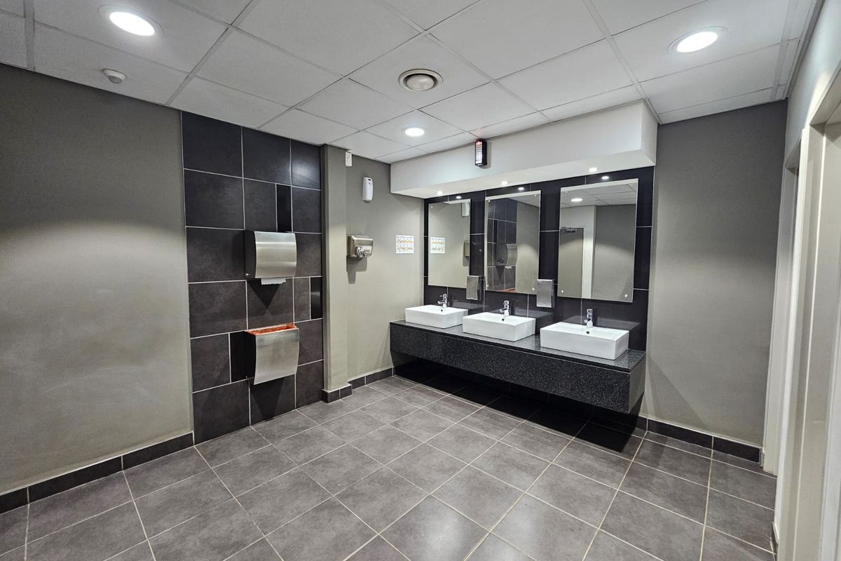 Modern commercial bathroom featuring a triple vanity with ceramic basins, granite countertop, wall-mounted mirrors, and professional tiled finishes.
