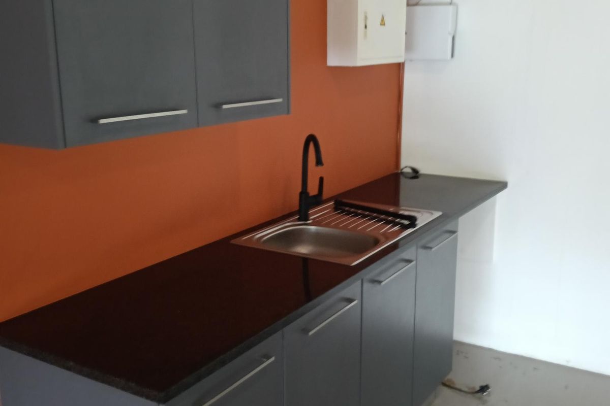 A compact office kitchenette featuring dark grey cabinetry, a black countertop, a stainless steel sink with a modern black faucet, and an orange accent wall.