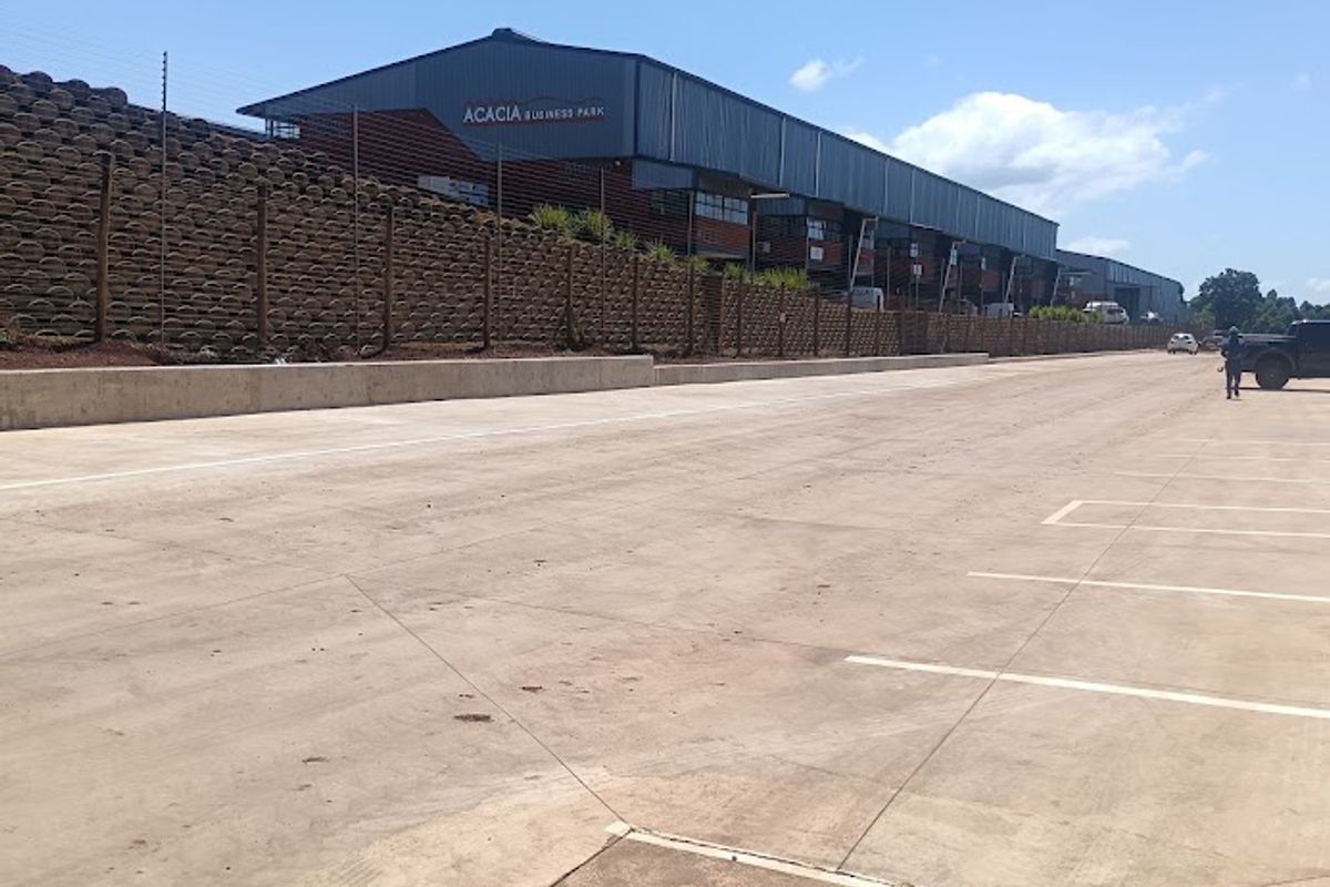 Exterior view of the Acacia Business Park industrial facility, featuring a large concrete parking area, retaining wall, and metal-clad warehouse structure.