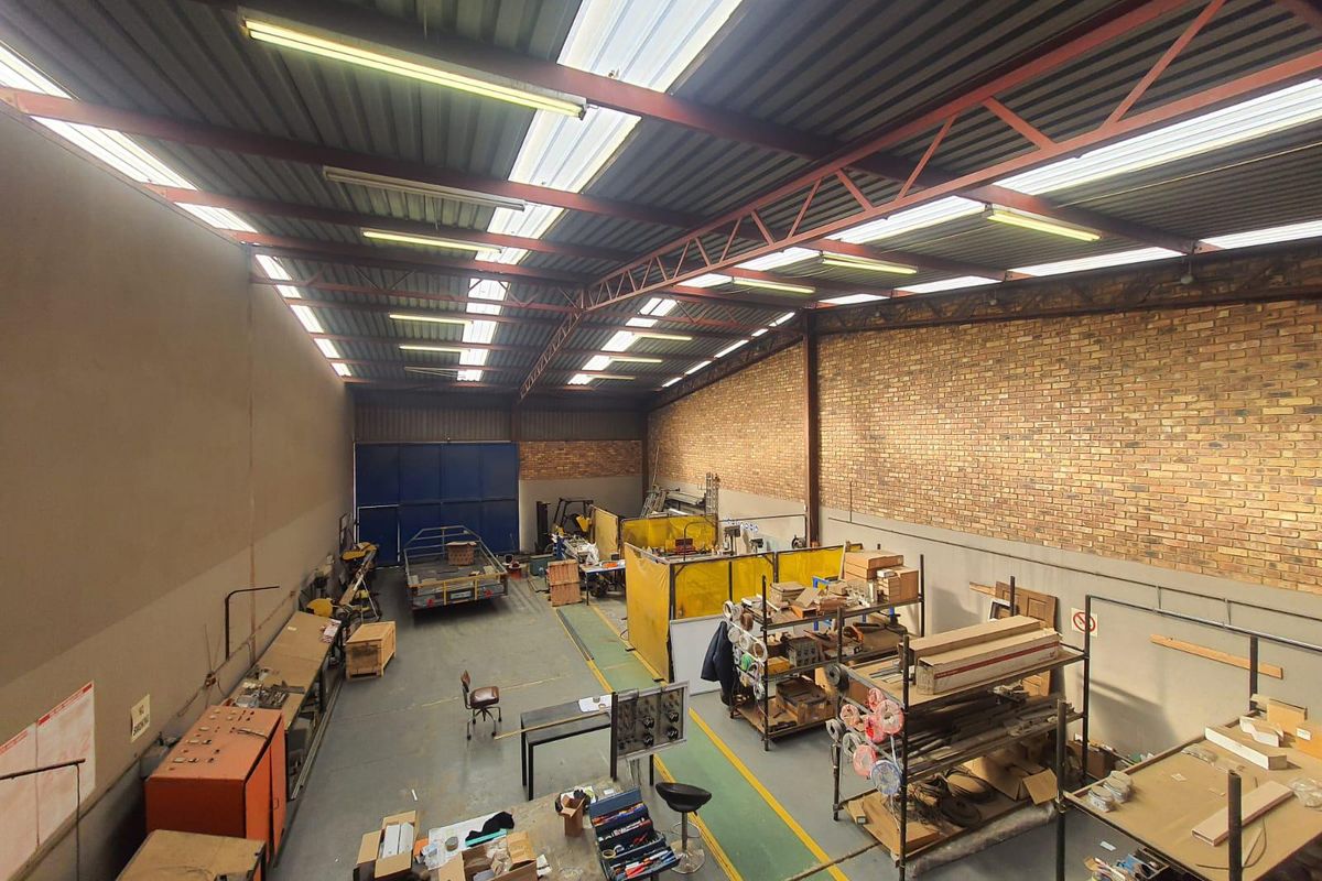 Spacious industrial warehouse featuring high-clearance steel truss roofing, translucent skylight panels, exposed brick walls, and large sliding access doors.