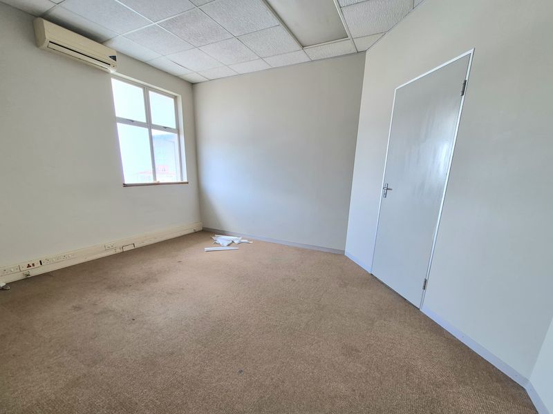 An empty office space featuring a suspended ceiling, wall-mounted air conditioning unit, and carpeted flooring with integrated electrical trunking.