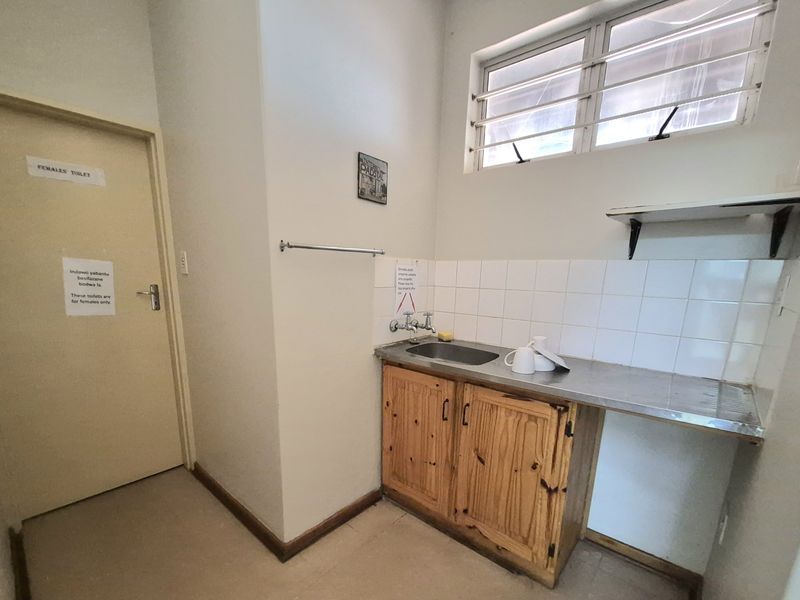 A functional office kitchenette featuring a stainless steel sink, tiled splashback, wooden base cabinetry, and wall-mounted shelving.