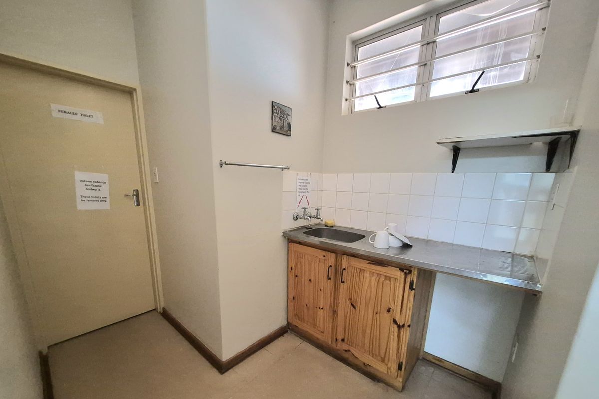 A functional office kitchenette featuring a stainless steel sink, tiled splashback, wooden base cabinetry, and wall-mounted shelving.