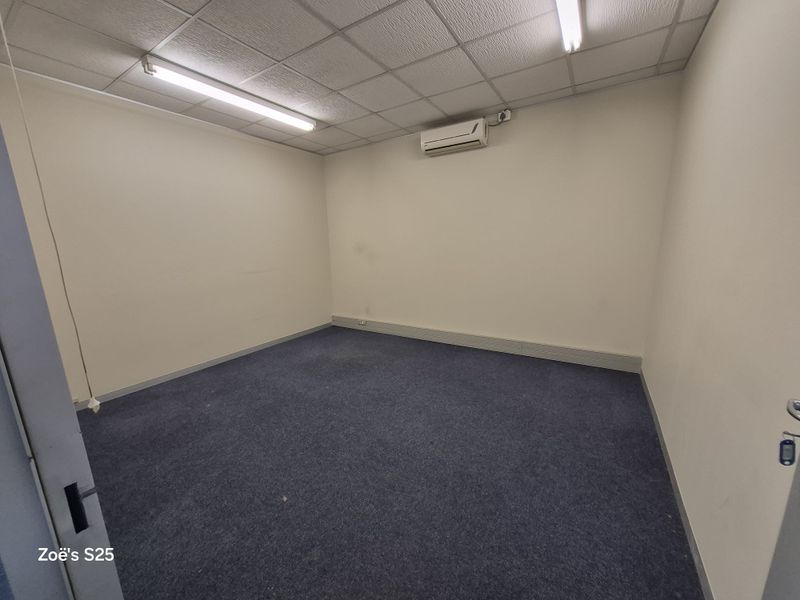 A vacant office space featuring a suspended ceiling, fluorescent lighting, a wall-mounted air conditioning unit, and blue carpeted flooring.