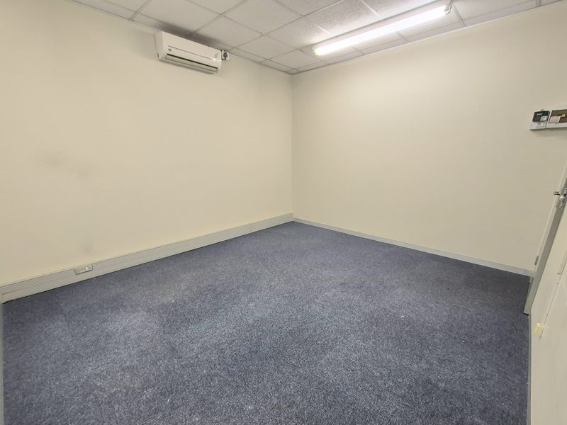 A vacant office space featuring carpeted flooring, a suspended ceiling with integrated lighting, and a wall-mounted split-system air conditioning unit.