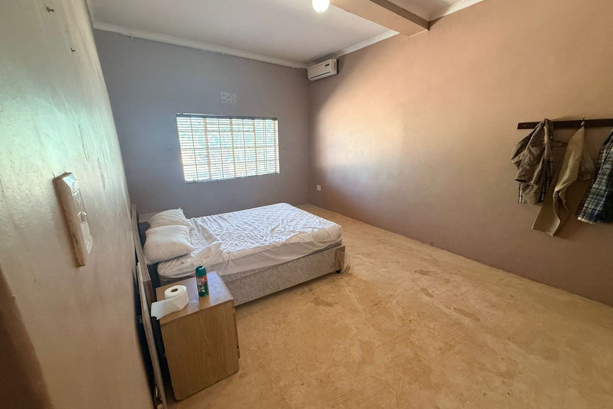 A spacious bedroom featuring a wall-mounted air conditioning unit, a window with blinds, and a simple, functional layout with concrete flooring.