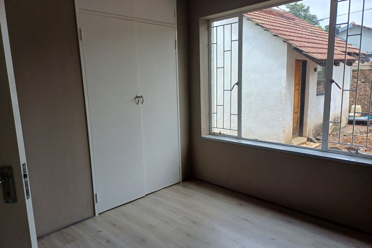 A bright, empty bedroom featuring light wood-look flooring, a built-in wardrobe, and large windows with security bars overlooking the rear exterior.