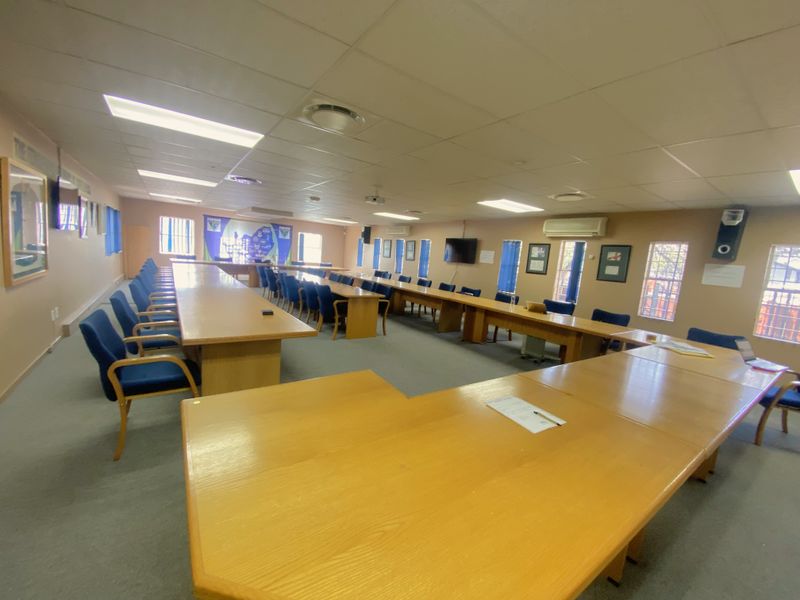 A spacious commercial boardroom or meeting area featuring built-in wooden tables, recessed ceiling lighting, and wall-mounted air conditioning units.