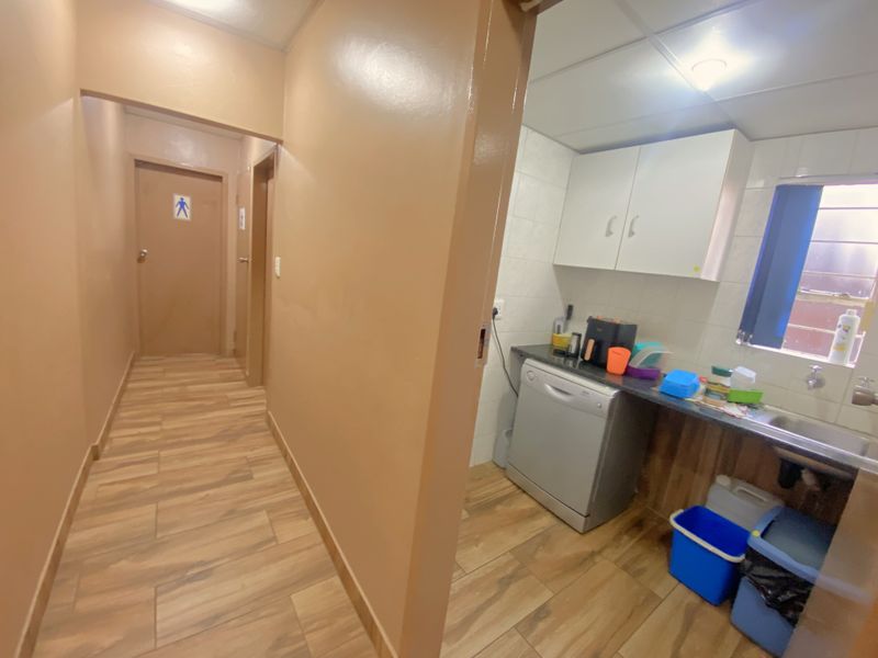A functional kitchenette area featuring built-in cabinetry, a sink, and tiled flooring, adjacent to a hallway leading to restroom facilities.